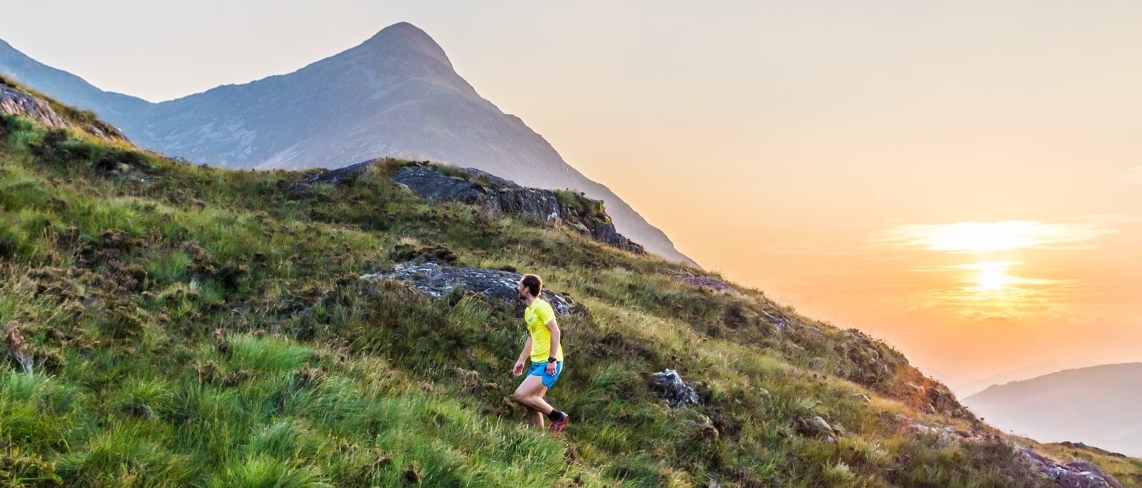 Trail runner ascending a grassy ridge above Loch Leven in the Scottish Highlands at sunset, with Pap of Glencoe rising behind