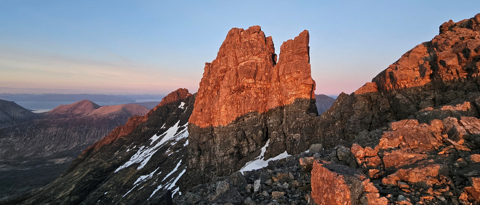 The twin-topped rock tower of the Bhasteir Tooth glows orange-red in low light, its lower half in cool shadow, with patches of snow on the slopes below and a calm sea and distant hills fading to pink on the horizon.