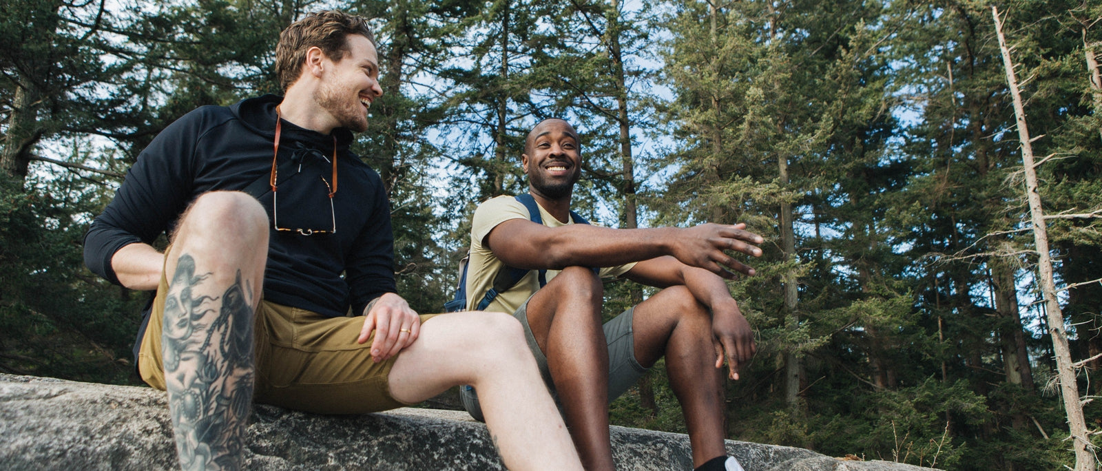 Two men sitting on a granite rock in a forest, laughing and relaxing in casual shorts.