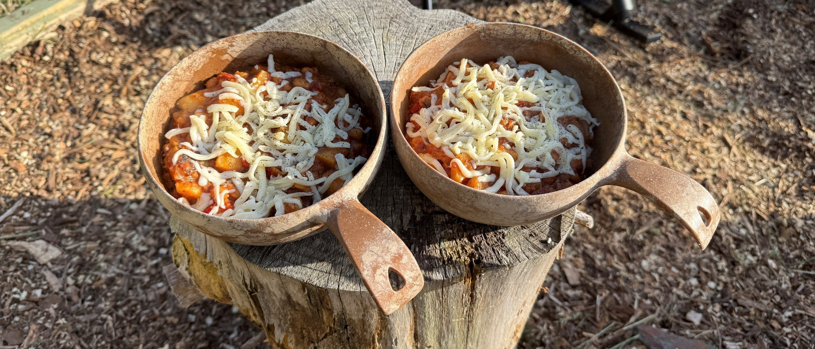 Two wooden camp bowls of corned beef hash topped with grated cheese, resting on a log stump outdoors