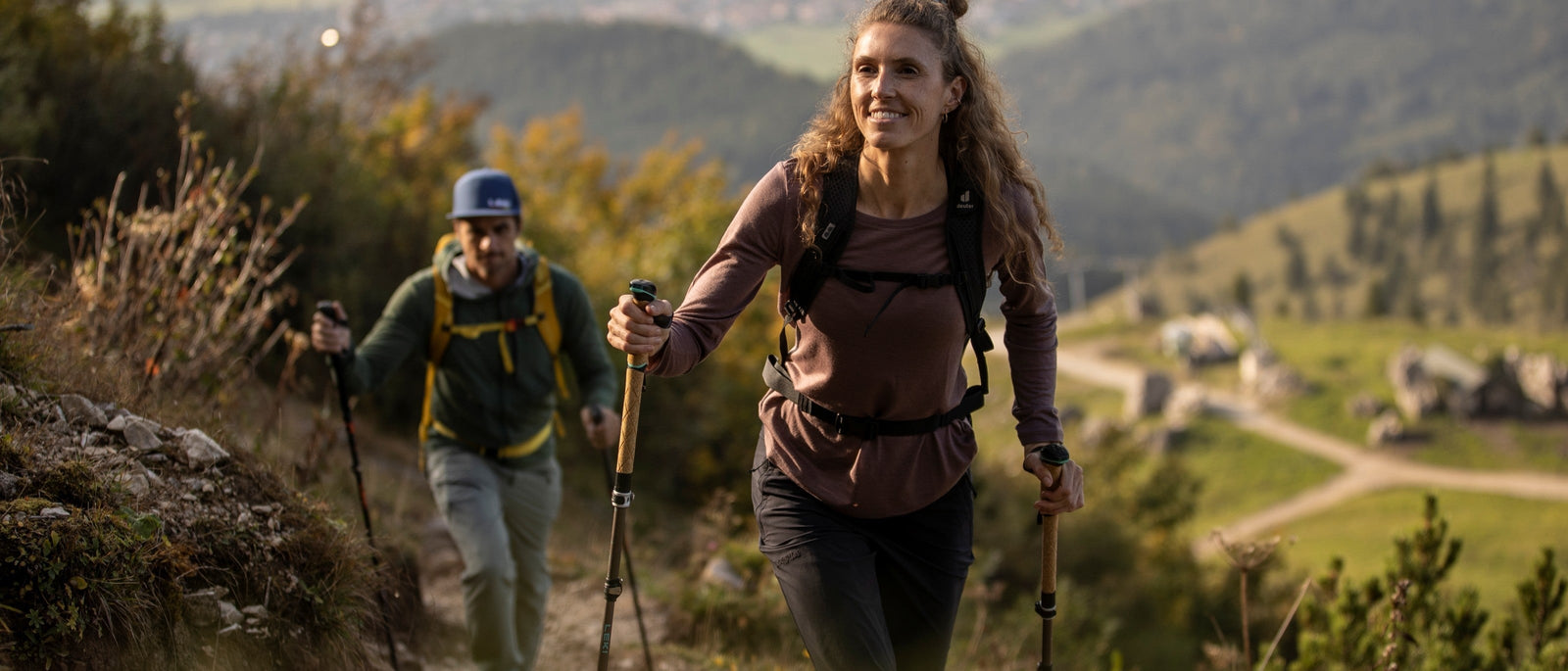 Two hikers ascending a grassy hillside trail using trekking poles for support on the climb.