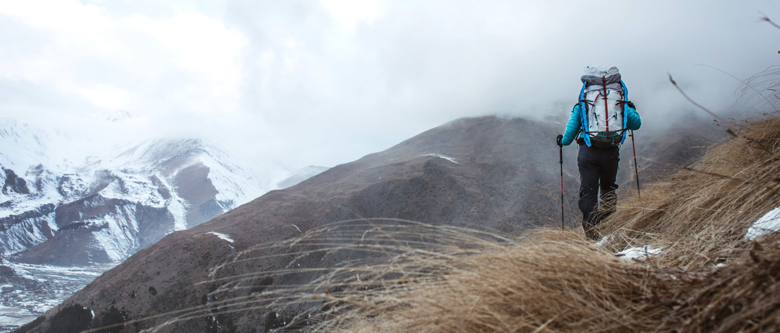 A backpacker in a teal down jacket climbs a slope with trekking poles, snow-streaked mountains rising beyond