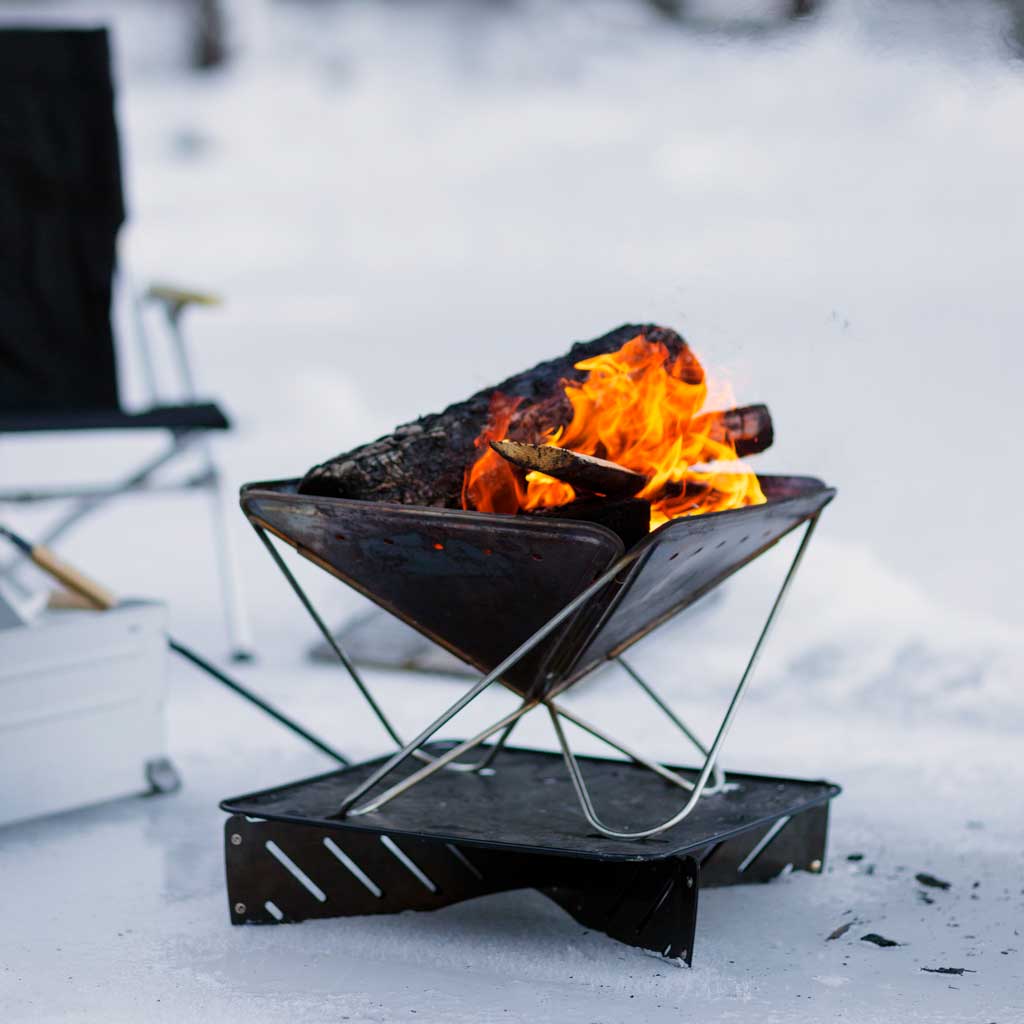 Snow Peak Fireplace portable fire pit burning with orange flames in snowy landscape, camping chair blurred in background