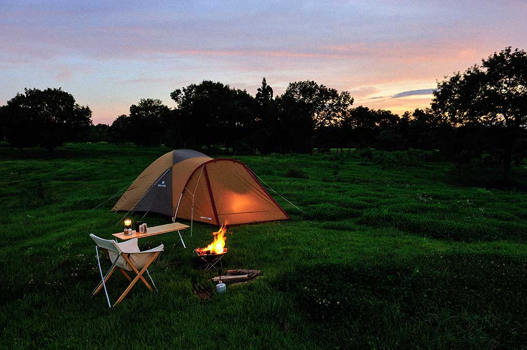 Illuminated tent with campfire, camping chair and table at dusk in a grassy field at sunset.