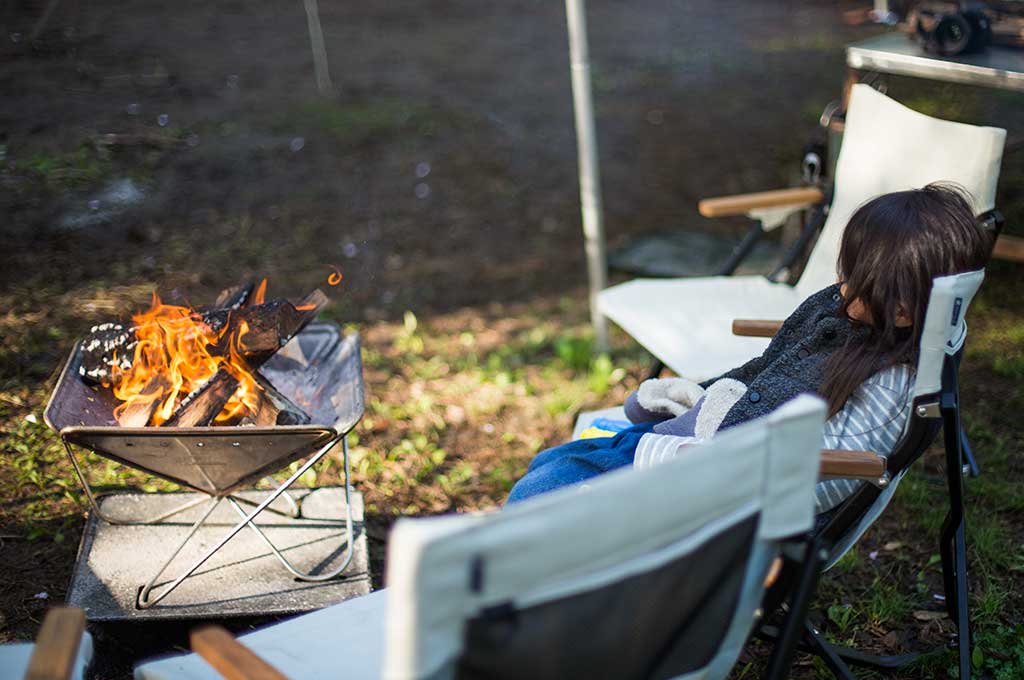 Person relaxing in a camping chair by a Snow Peak Pack & Carry Firepit with flames in a garden setting.