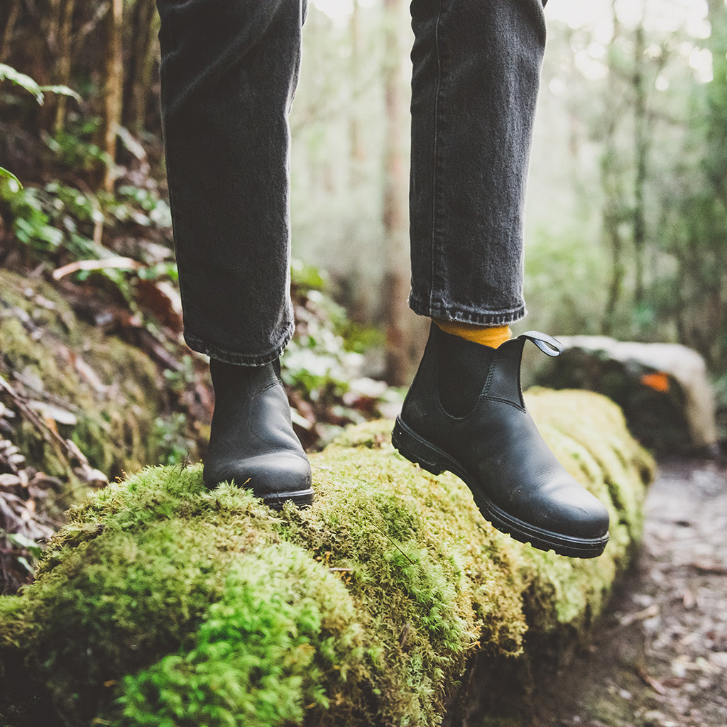 Person wearing black Chelsea boots and jeans standing on a moss-covered log in a woodland setting.