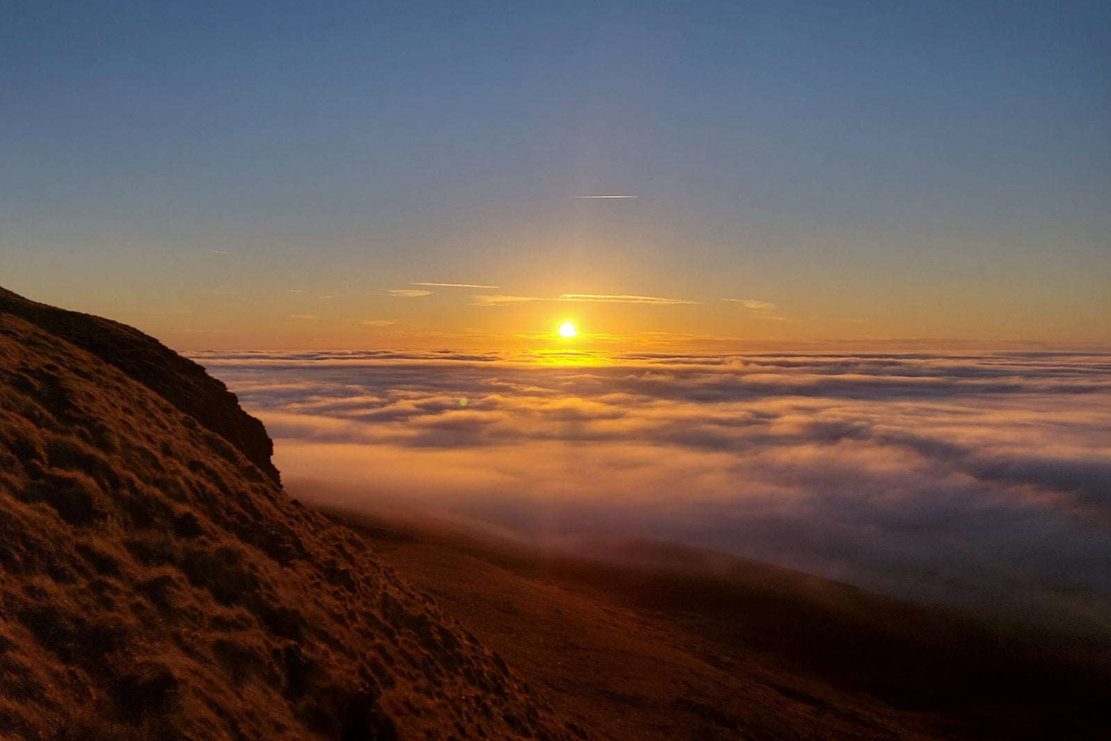 Sunset from Pen-y-Ghent with golden sun above a sea of clouds and rocky mountainside in foreground.
