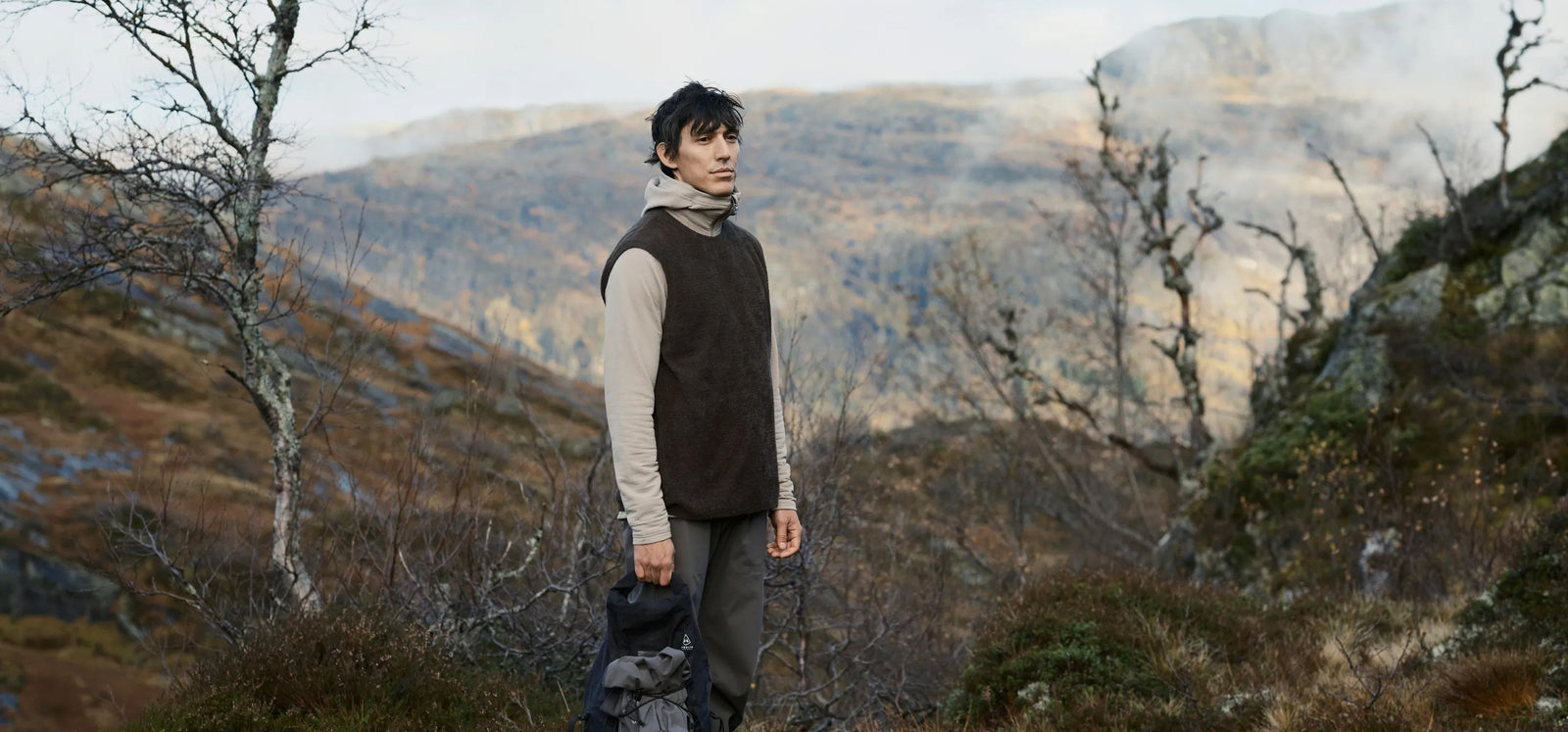 Person wearing Houdini outdoor clothing standing on a hillside with mountain views in background.