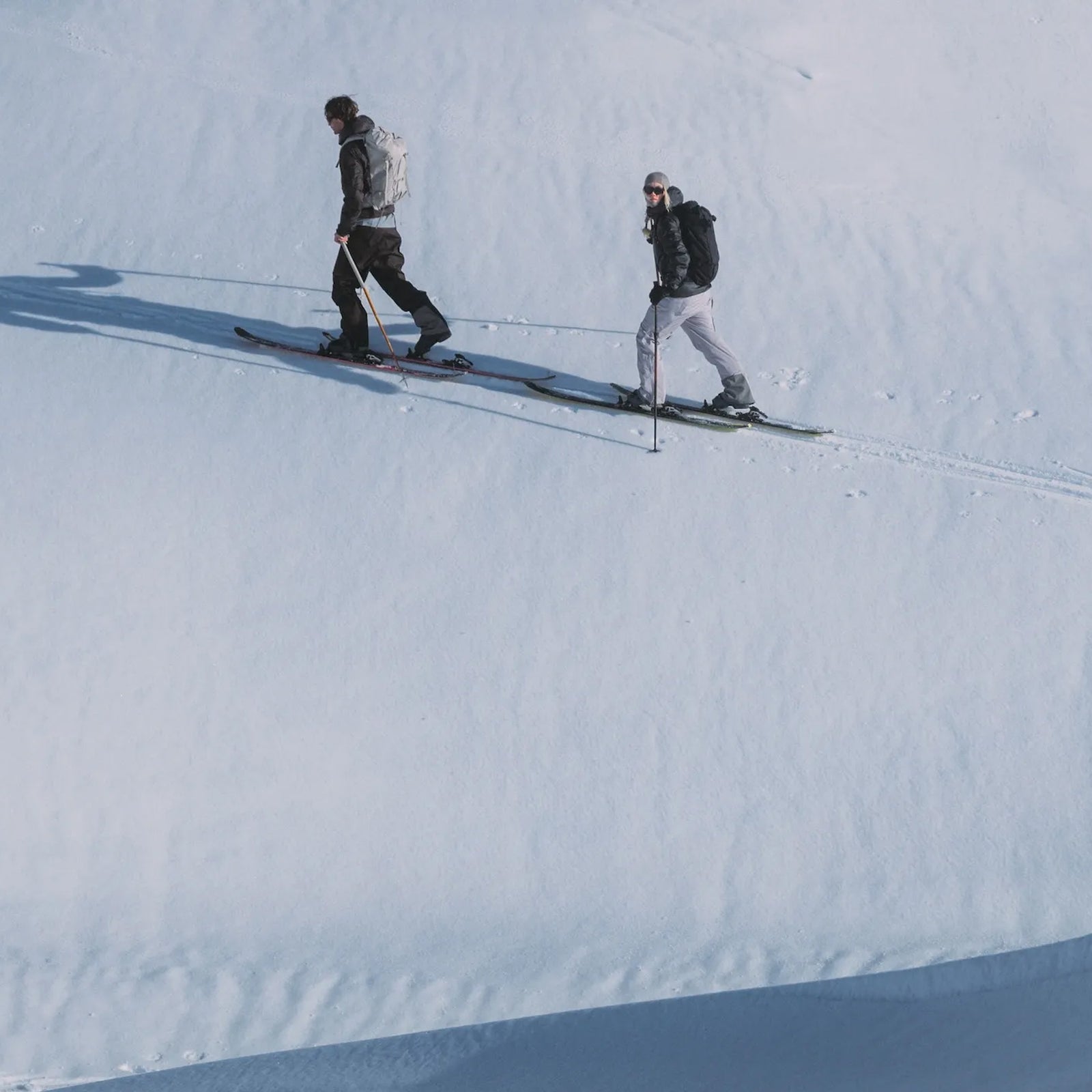 Two skiers cross-country skiing across snowy terrain in winter clothing and backpacks.