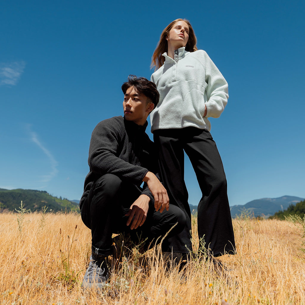 Two people wearing Columbia fleeces in a sunny field with mountains in the background.