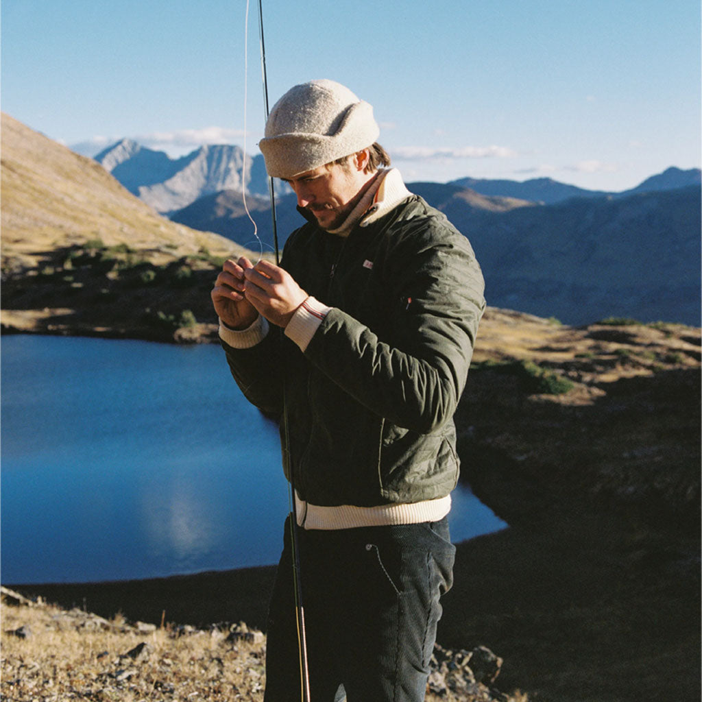A person fly fishing by an alpine lake in mountains, wearing Amundsen Sports outdoor gear.