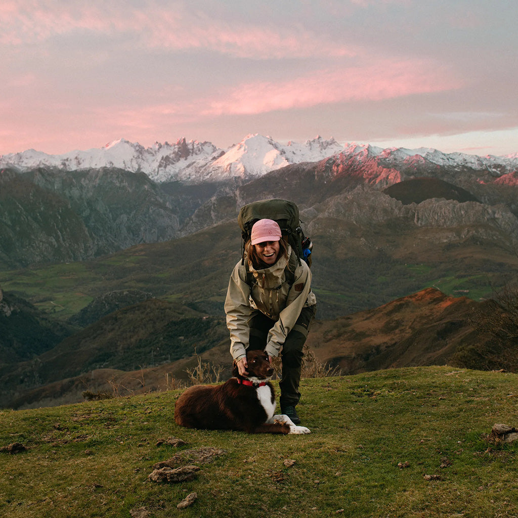 A hiker with a backpack and dog pose on a mountain ridge at sunset, snow-capped peaks in the distance.