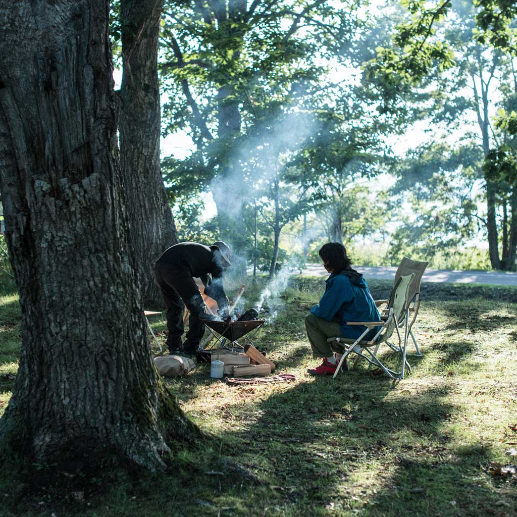 Two campers tending a campfire under tall trees in a woodland clearing.