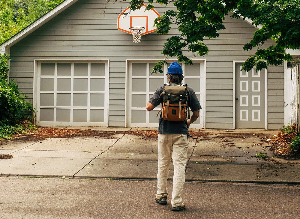 A hiker wearing a brown canvas backpack and blue baseball cap runs towards a house with a basketball hoop.