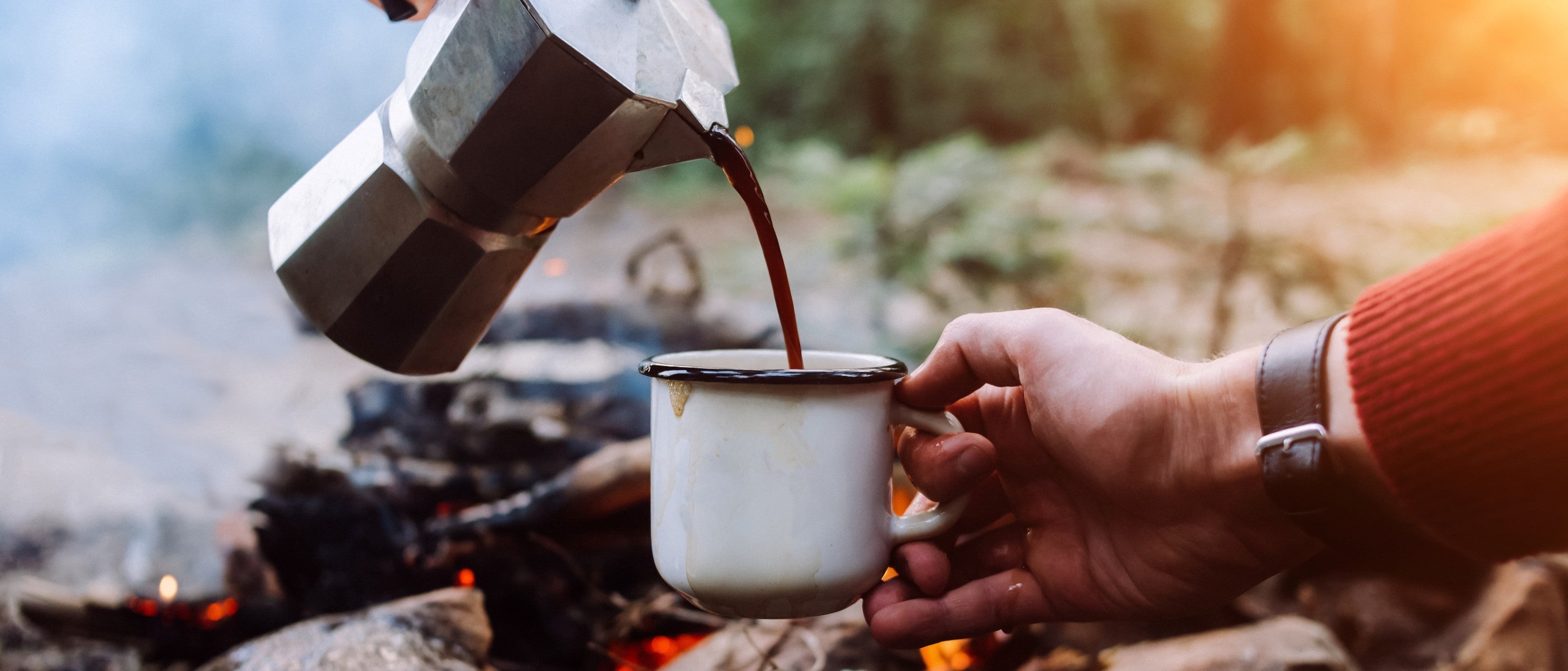 Freshly brewed coffee being poured from a moka pot into a camping mug beside a roaring campfire.