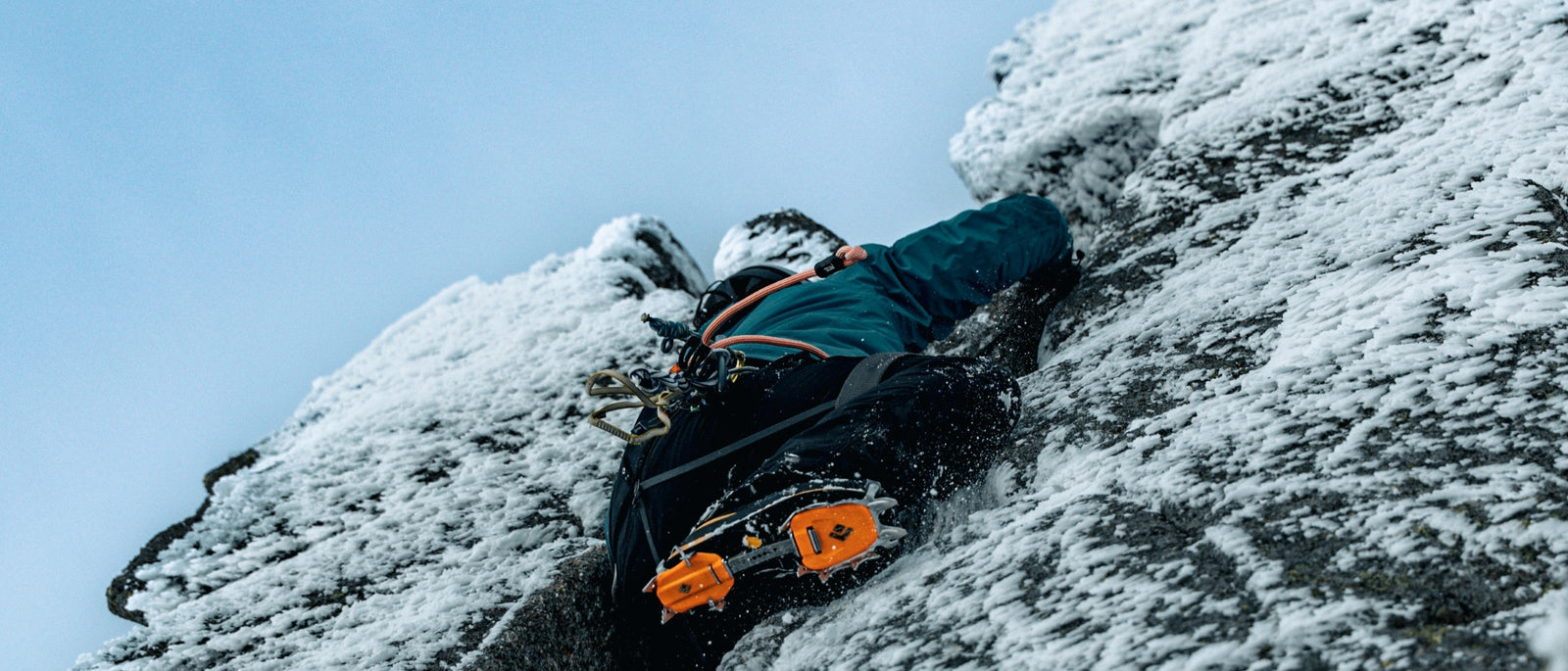 A climber in crampons stemming up a snow-encrusted rock chimney on a Scottish winter crag, viewed from below against a pale sky.
