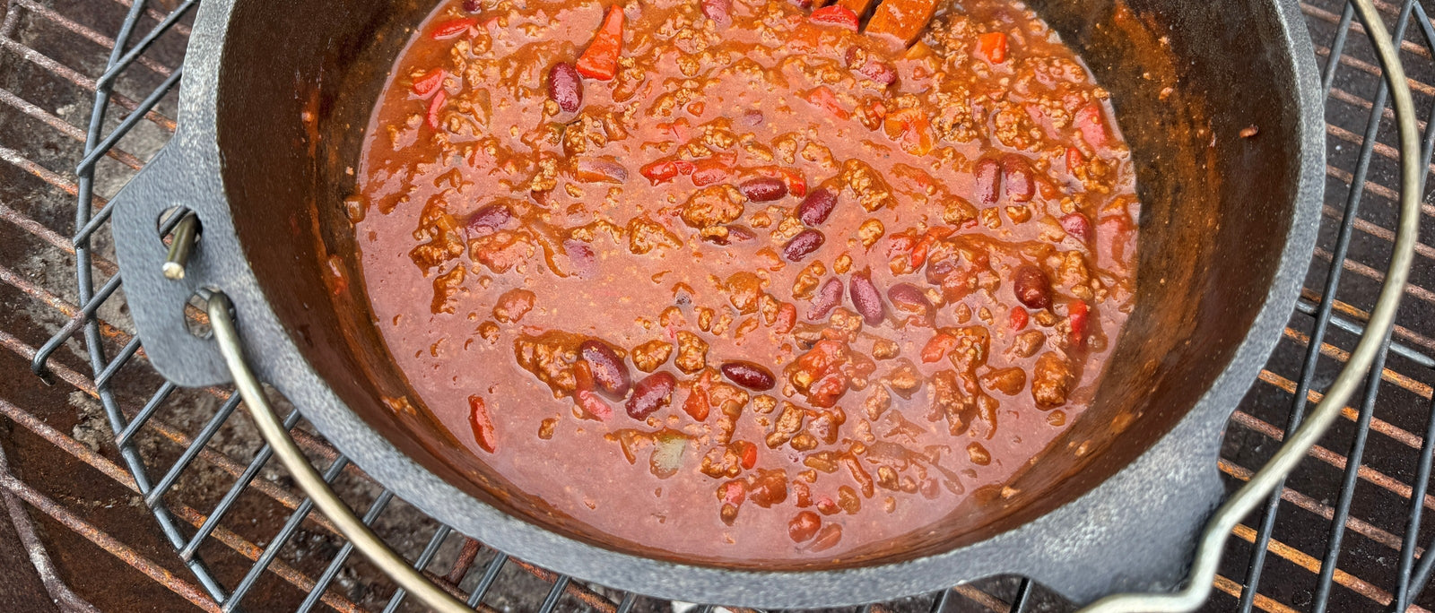 Close-up of chilli con carne simmering in a cast iron Dutch oven over a campfire grill, with kidney beans, beef mince and red pepper visible in a rich tomato sauce