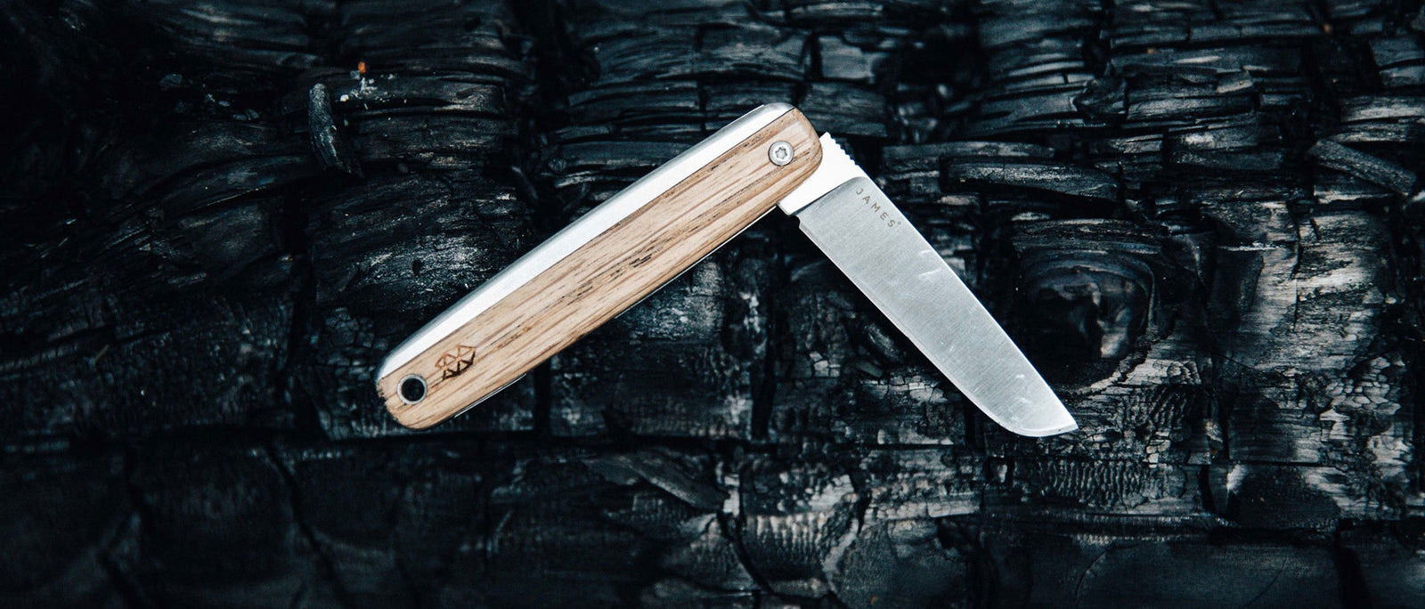 Wooden-handled folding pocket knife by The James Brand with a straight blade, partly open on a charred log.