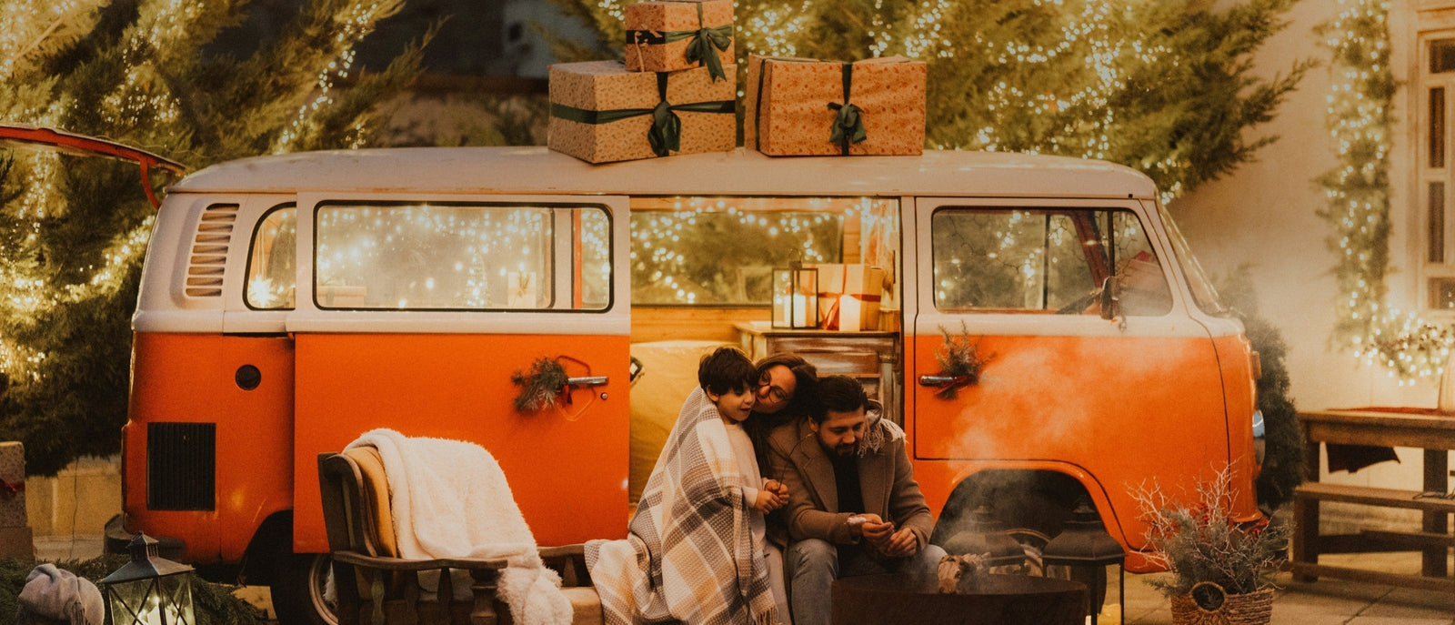 Festive scene of outdoorsy family sitting outside their vintage VW camper van surrounded by gifts and fairy lights.