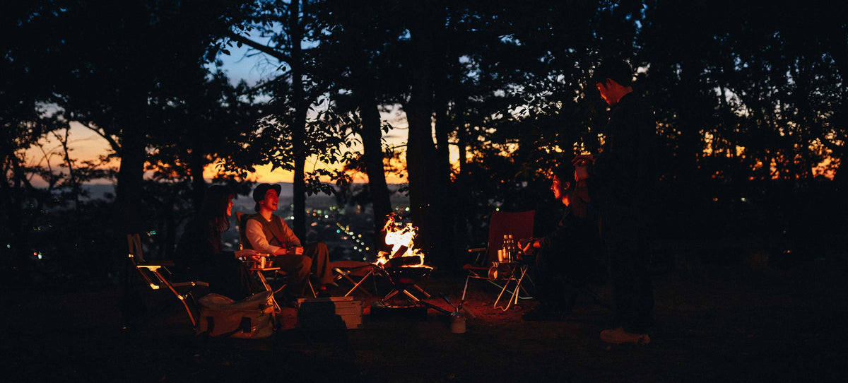 Campers gathered around a glowing campfire at dusk under tall trees, with a golden sunset sky visible beyond.