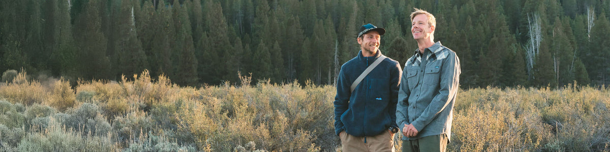 Two men wearing autumn outdoor clothing in a sagebrush landscape with coniferous forest behind.