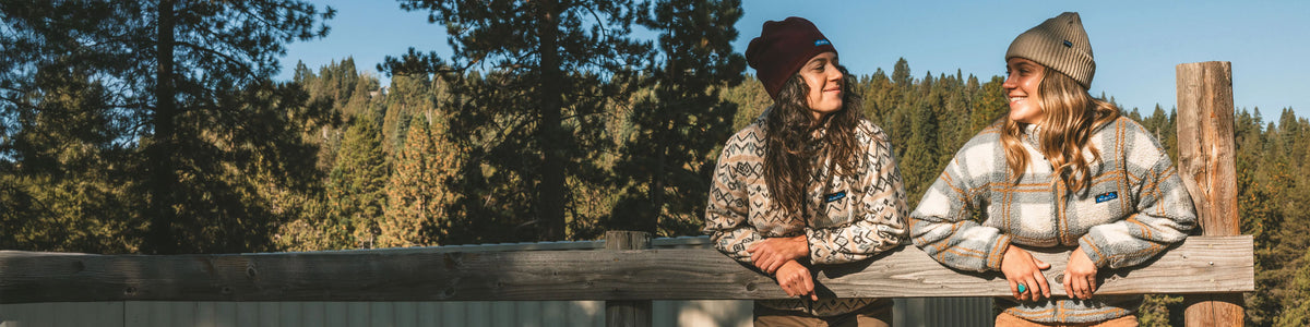 Two women wearing autumn clothing and beanies sit on a wooden rail overlooking a coniferous forest in sunlight.