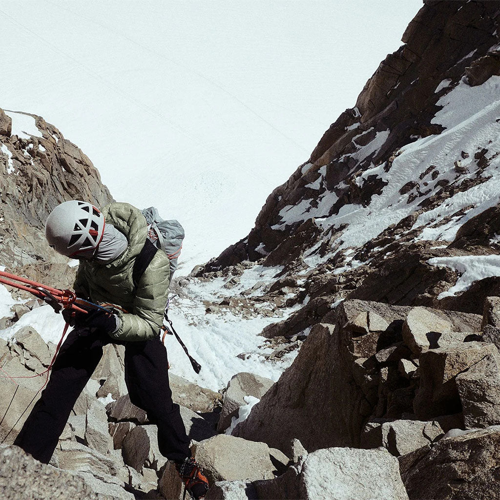 A climber wearing a white helmet and olive jacket navigates rocky, snow-covered alpine terrain on a mountain face.