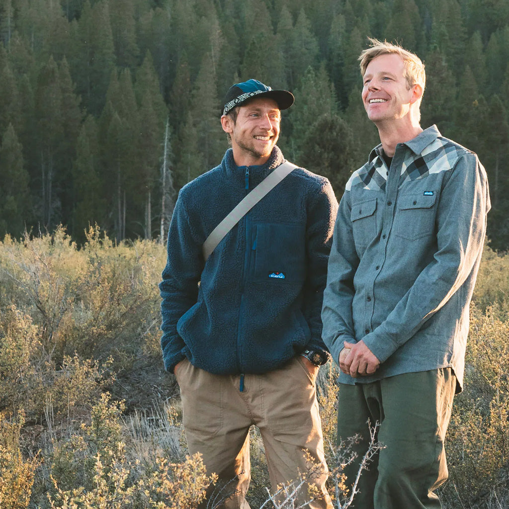 Two hikers in blue fleeces and shirts stand in a sunlit forest clearing with coniferous trees behind them.