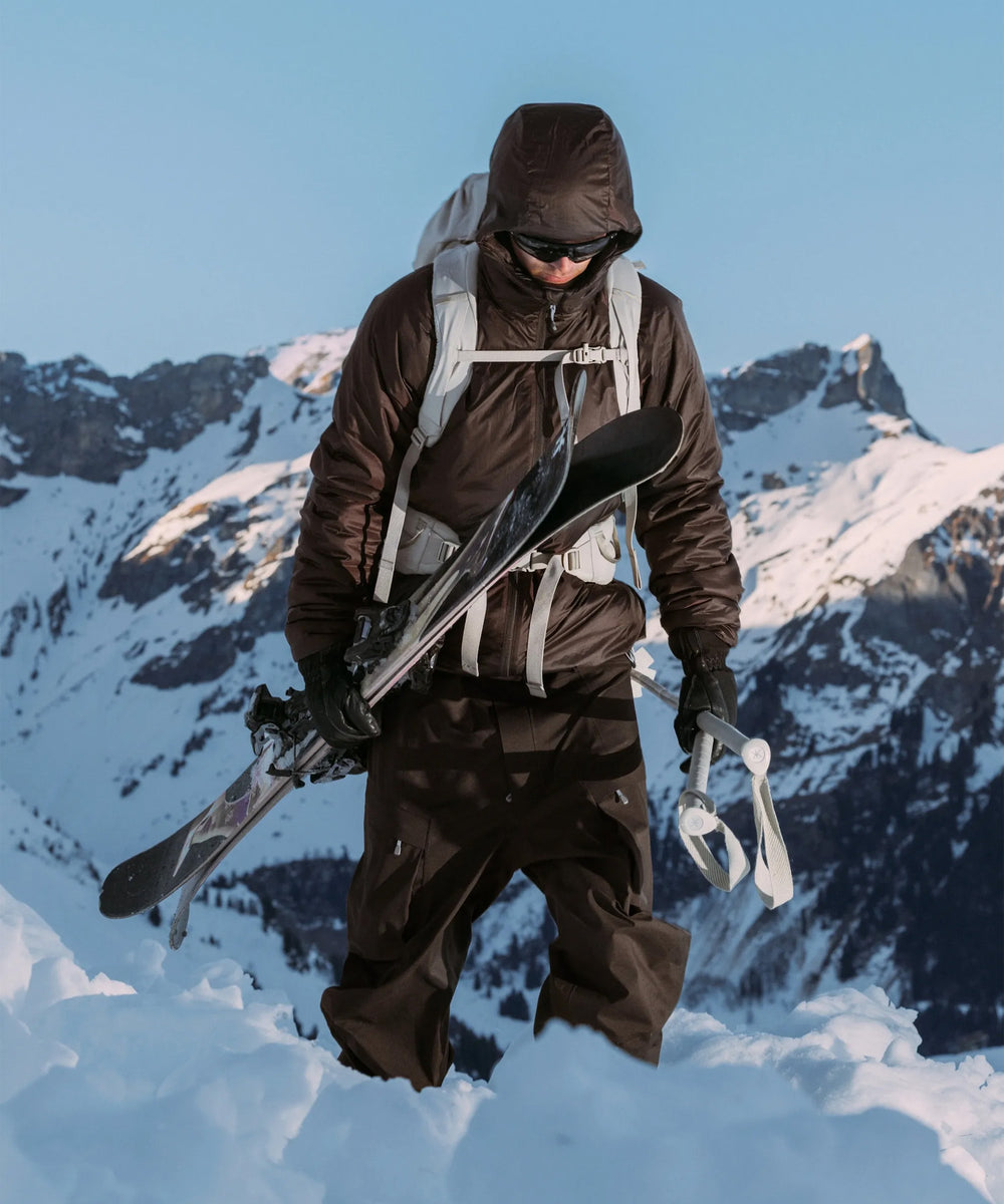 Skier in brown Houdini jacket and backpack carrying skis in snowy mountain landscape.