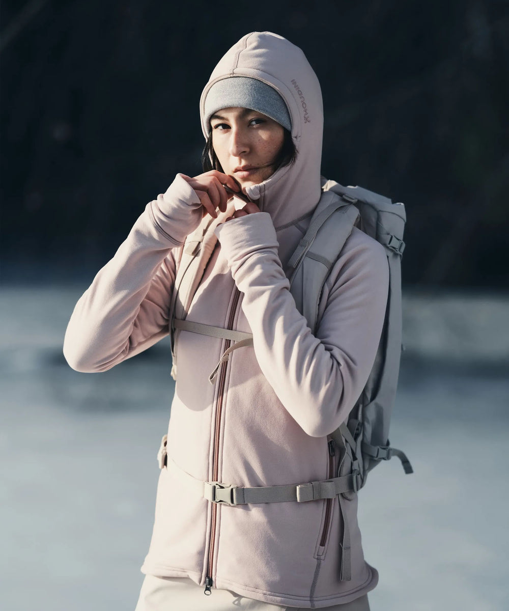 Woman wearing Houdini pale pink hooded jacket and matching beanie with backpack near water.