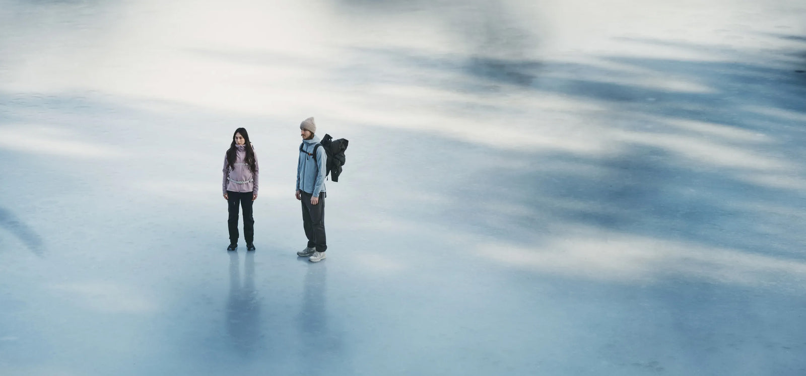 Two people standing on a frozen lake wearing Houdini outdoor gear, one holding a jacket.