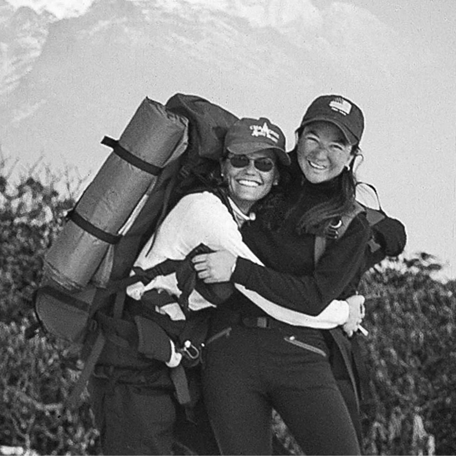 Two hikers smiling at the camera in mountainous terrain, one carrying a rolled sleeping bag.