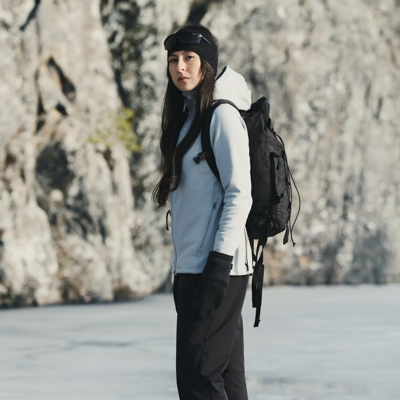 Woman wearing Houdini Houdi series outdoor gear with black backpack against a rocky cliff face.