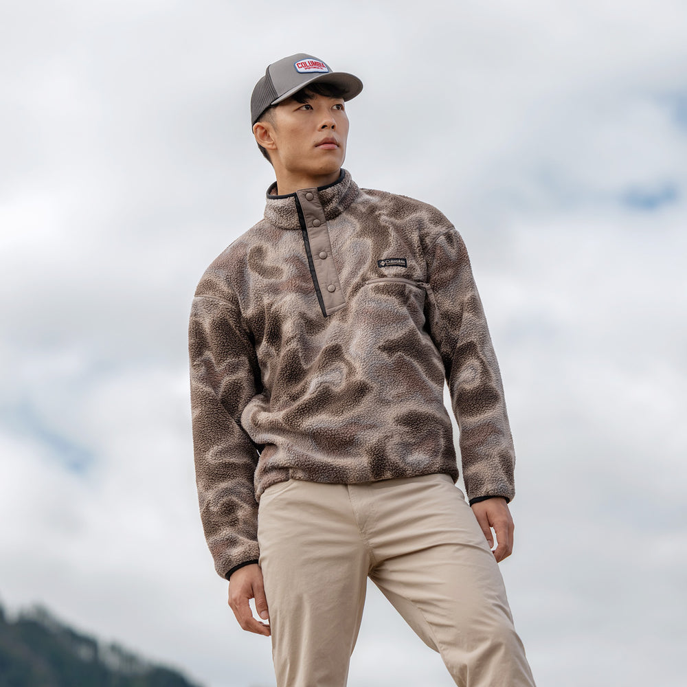 Person wearing a grey Columbia fleece pullover and beige trousers with a grey cap against a cloudy mountain backdrop.