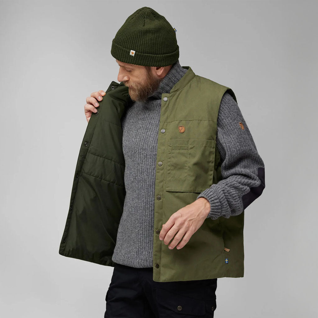 Man wearing a Fjällräven khaki-green padded gilet over a grey knit sweater, styled with a green beanie and black trousers.