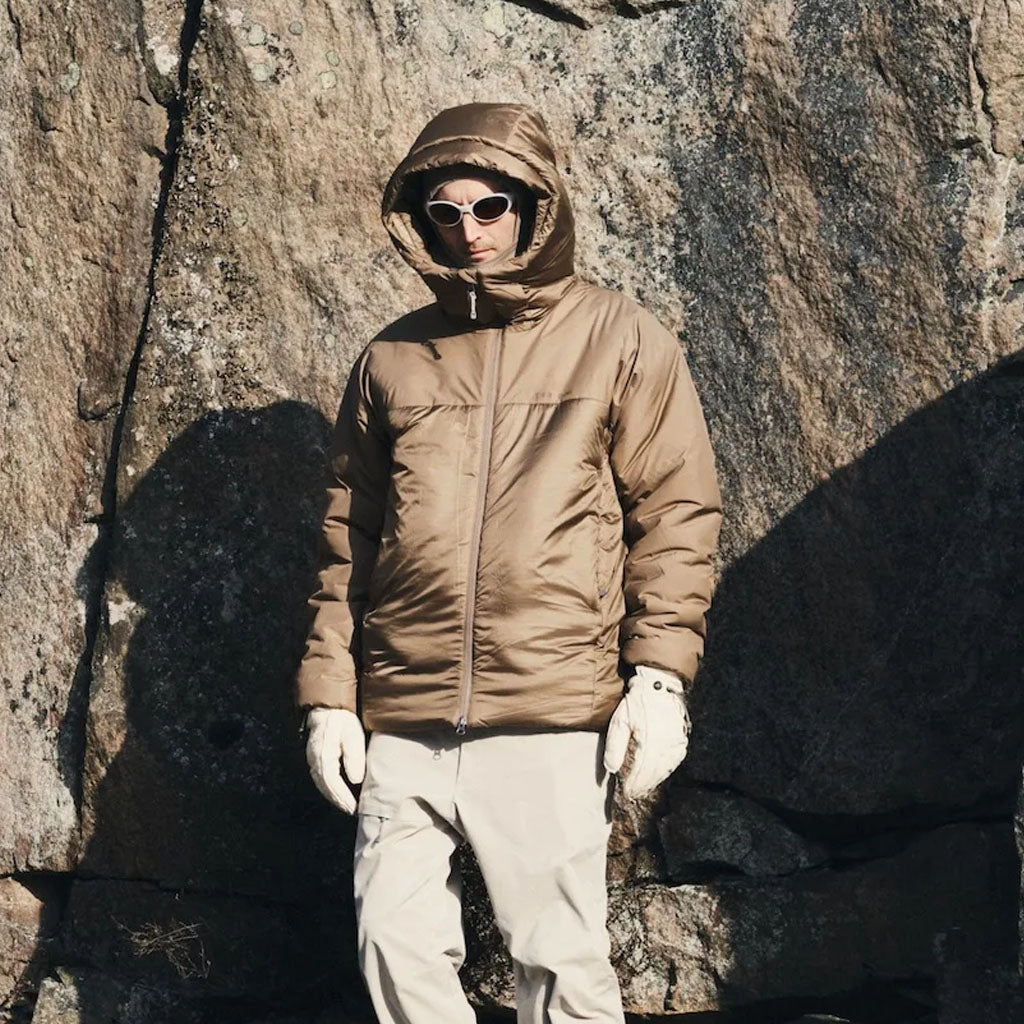 Person wearing beige Houdini puffer jacket with hood, white trousers and sunglasses against a stone wall.