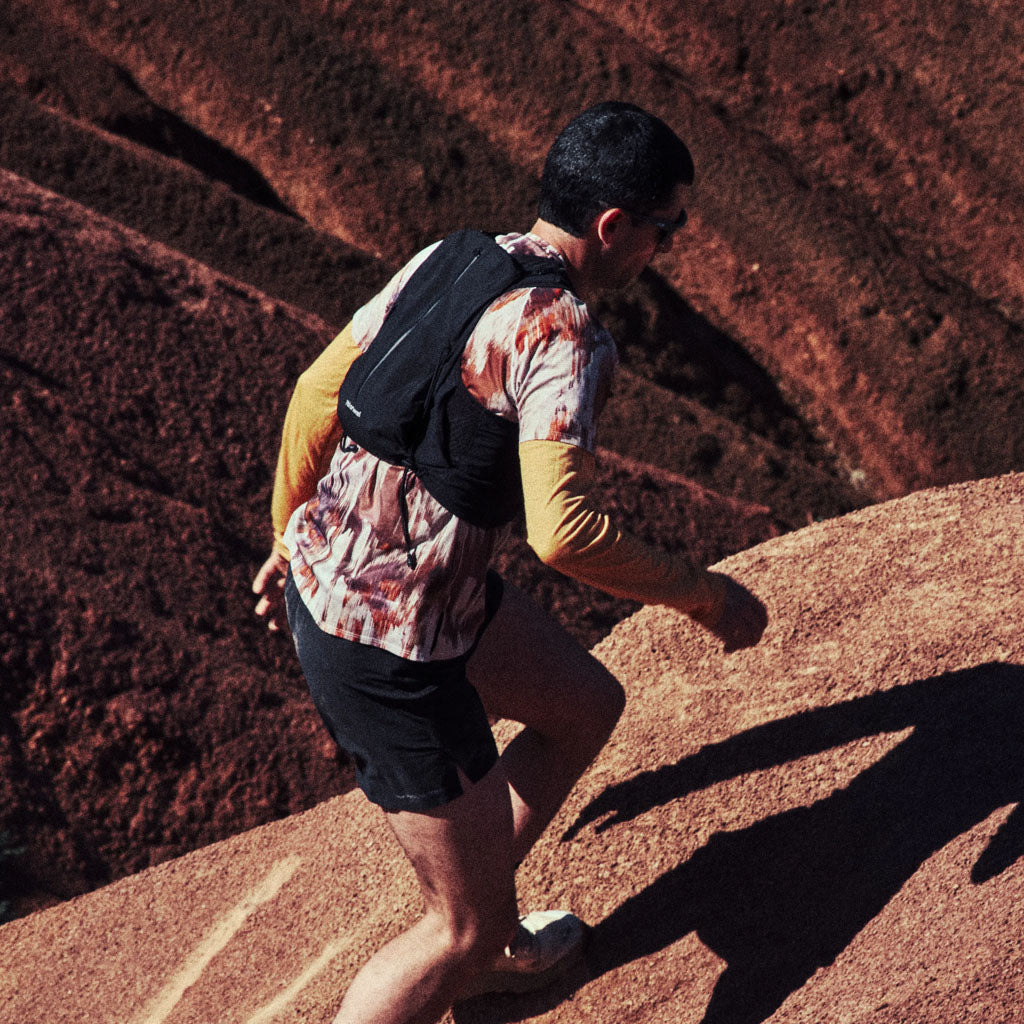 A hiker wearing a black backpack and patterned jacket runs up a steep, rocky mountain slope in bright sunlight.