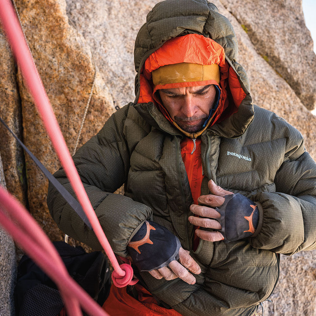 A climber in a Patagonia black insulated jacket and red hood secures a rope against a granite rock face.