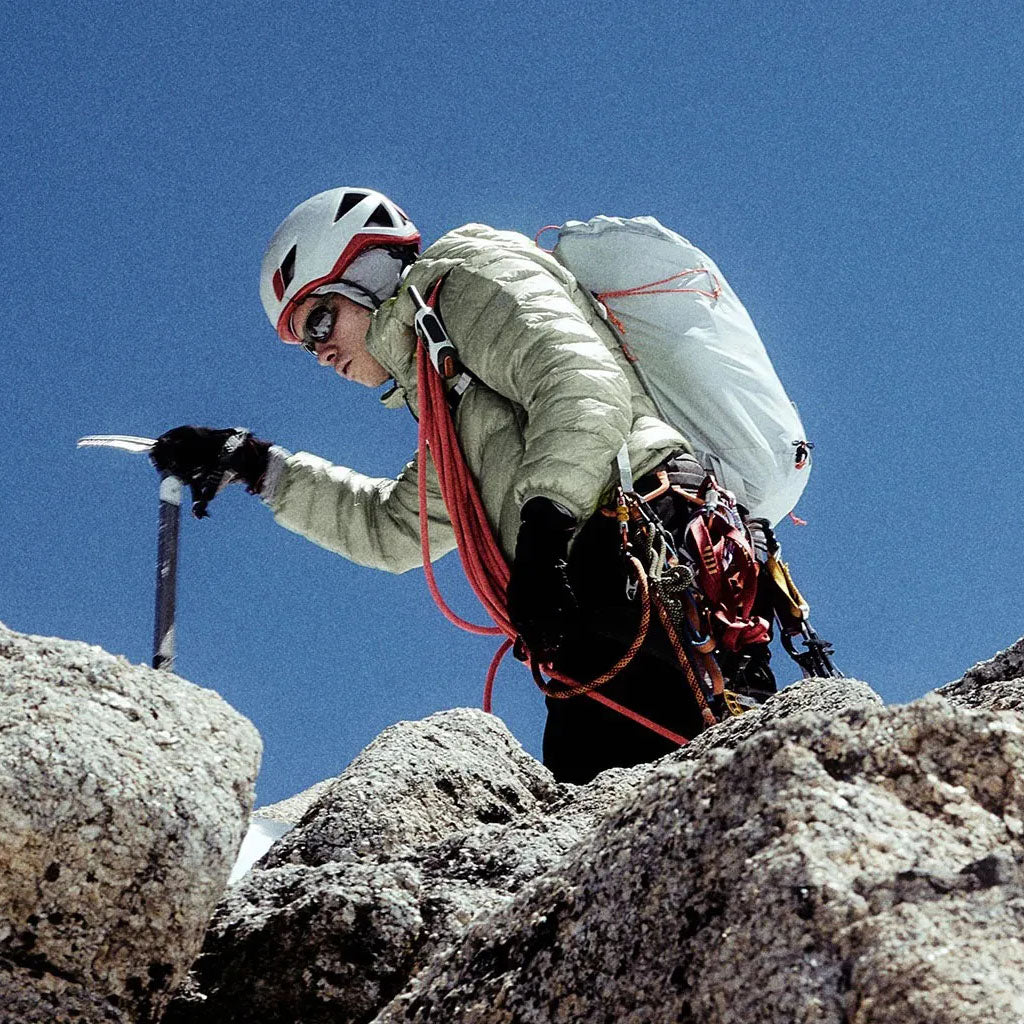 A climber in helmet and harness ascending a rocky mountain against a clear blue sky.