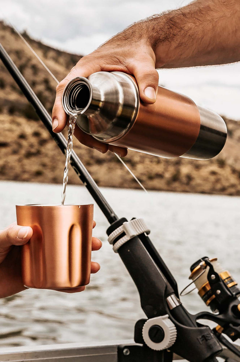 Person pouring water from a copper thermos into a copper cup while fishing by a river.