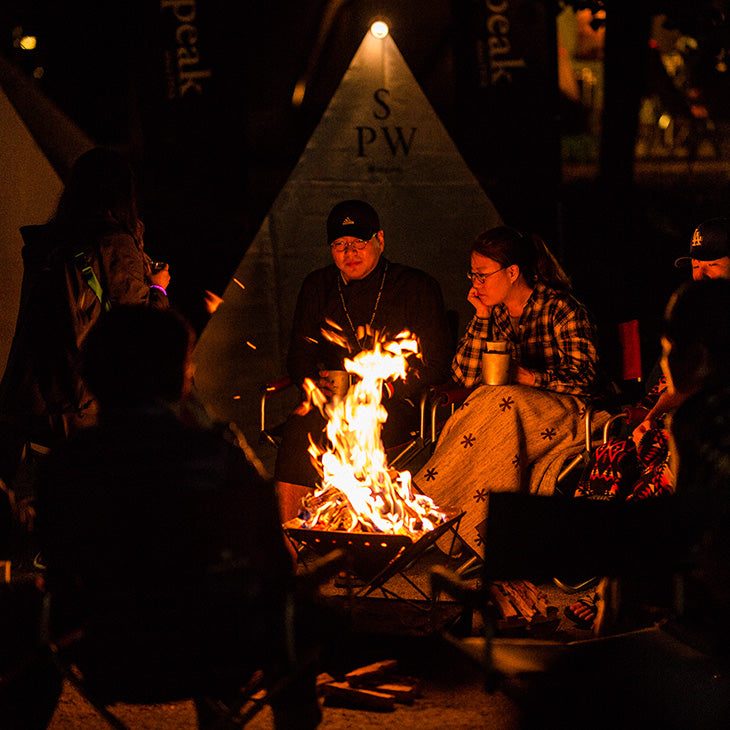 People gathered around a campfire at night beside a canvas bell tent, warming themselves and socialising in the outdoors.