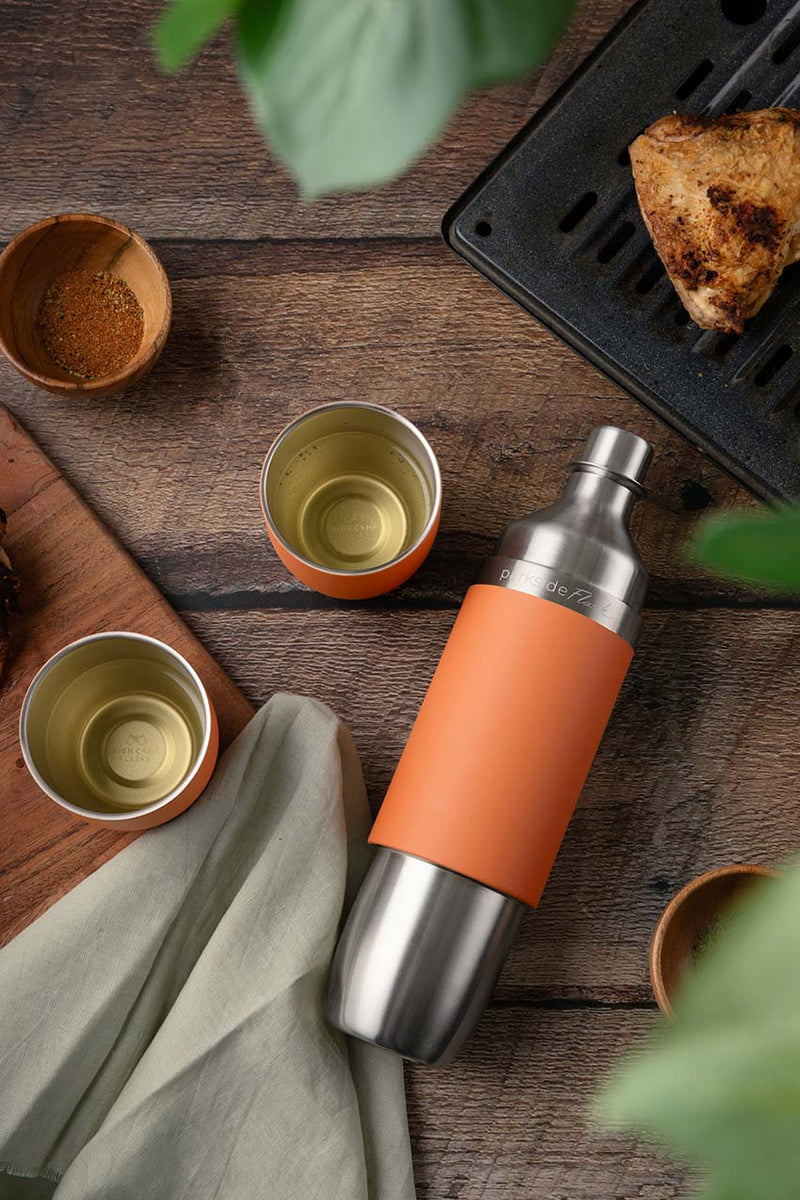Parkside orange insulated water bottle with cups of tea, spices and camping food on a wooden surface.