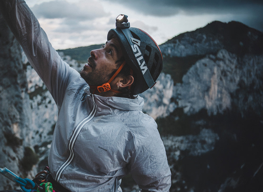 A climber wearing a Silva headtorch and white jacket looks up at a dramatic rocky cliff face.