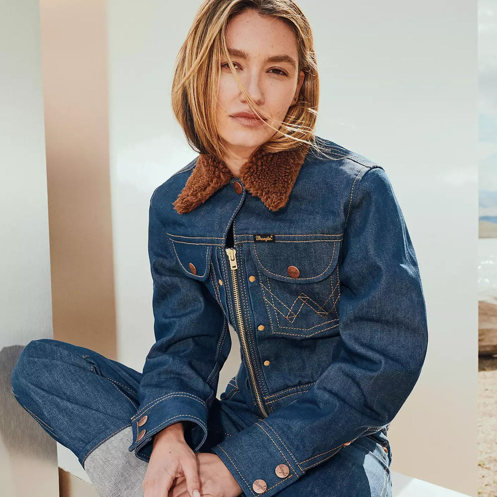 Woman wearing Wrangler dark denim jacket with brown sherpa collar and matching jeans, seated indoors.